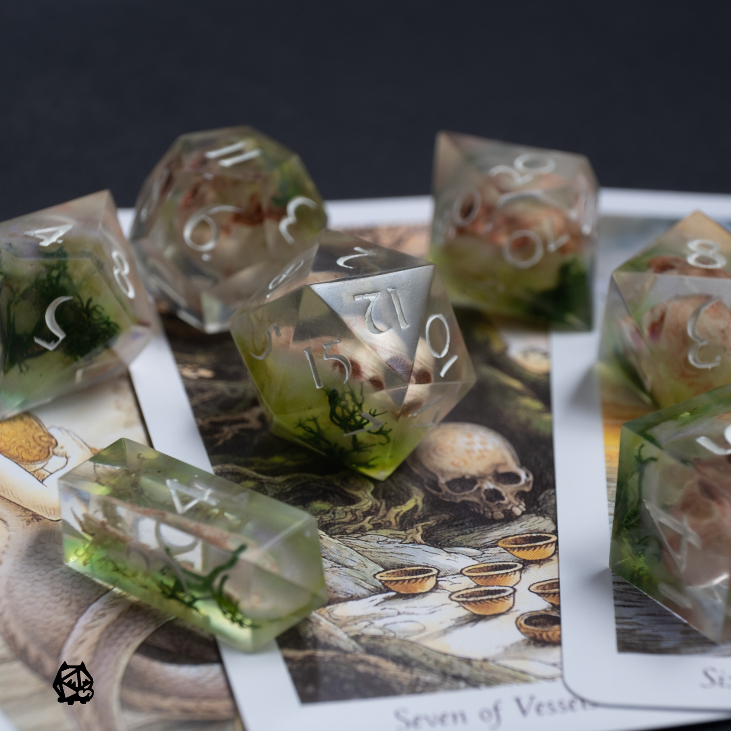 A set of clear resin polyhedral dice with hand painted skulls, displayed on a tabletop with game-related artwork.